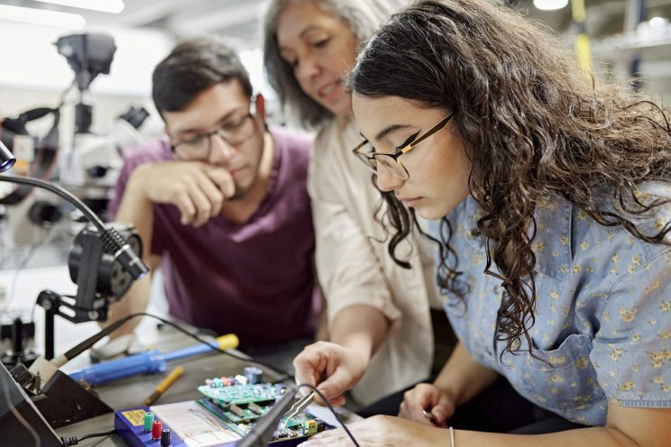 Photograph of a teacher showing two students how to connect circuits on a circuit board.
